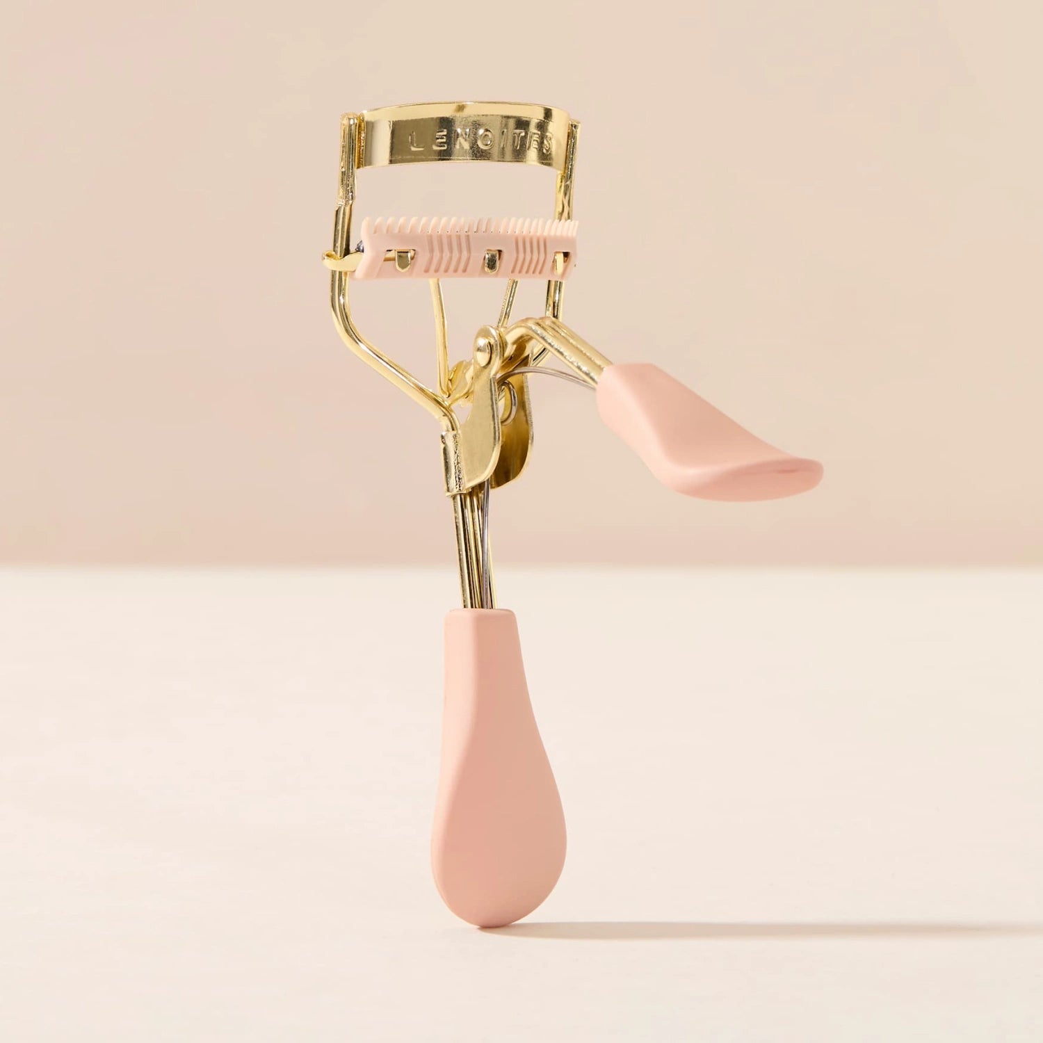 Eyelash Curler Lash Lift by Lenoites, featuring a pink eyelash curler with gold details and a black rubber pad, accompanied by a small pink comb, displayed against a soft beige background.